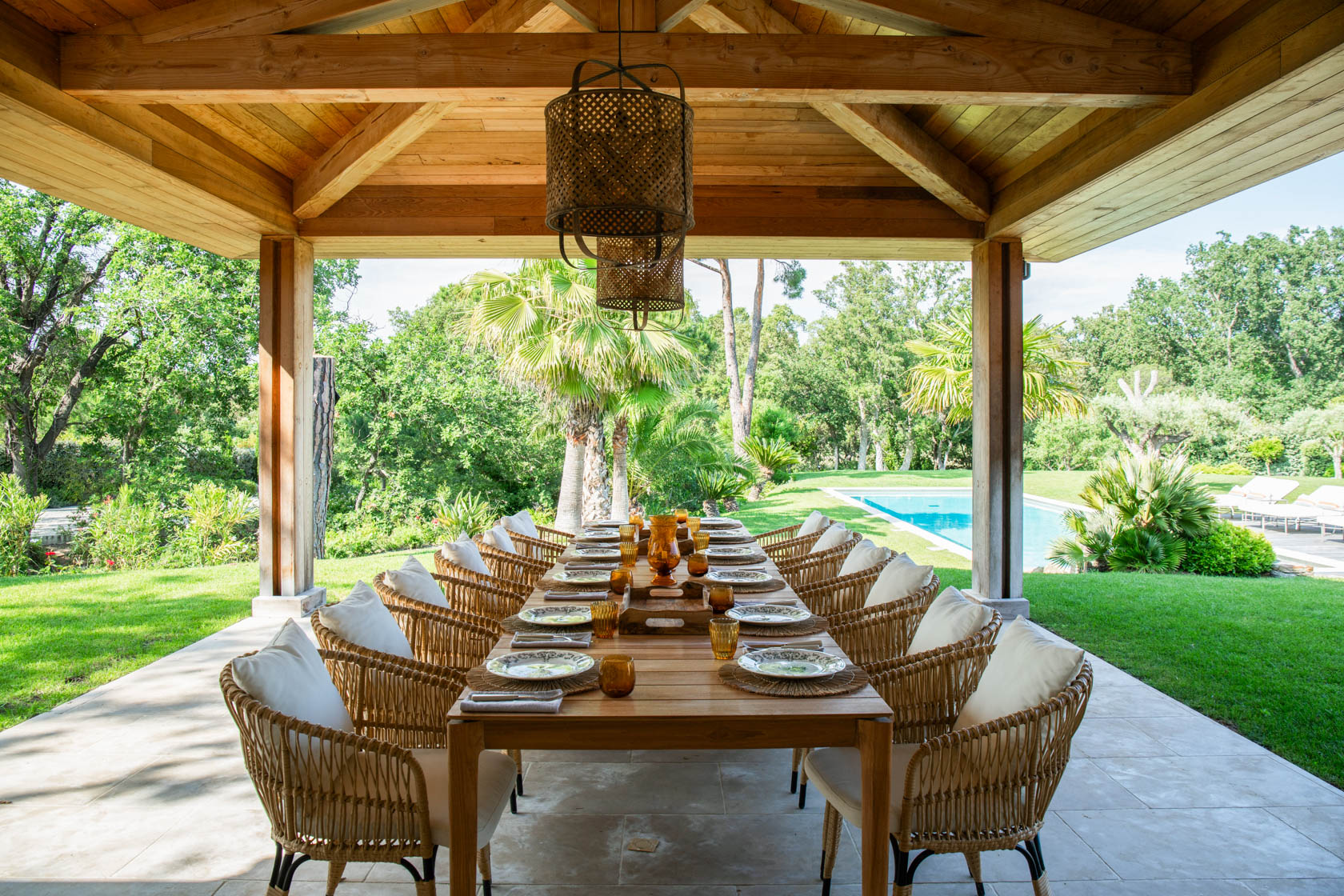 grande-table-a-manger-en-bois-entouree-de-chaises-en-osier-avec-des-coussins-blancs-sous-une-veranda-et-deux-luminaires-dans-un-jardin-avec-piscine
