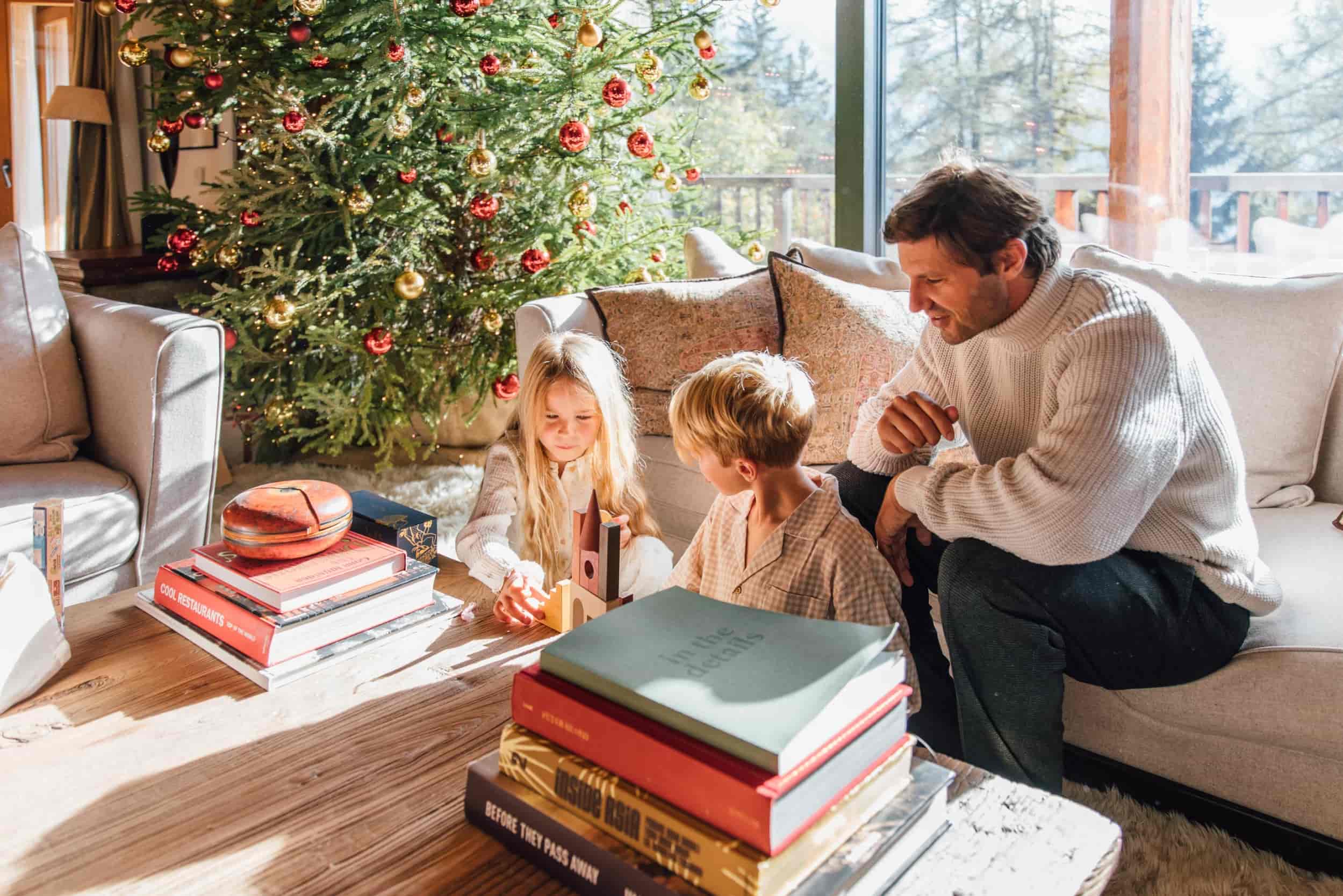 A-father-sad-on-a-coach-next-to-a-decorated-christmas-tree-watching-his-blond-son-and-daughter-play-with-wooden-toys-on-a table-covered-in-books