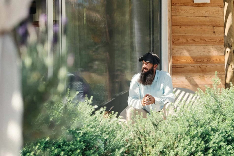 homme-brun-avec-un-beret-devant-une-maison-en-bois-entouree-de-verdure