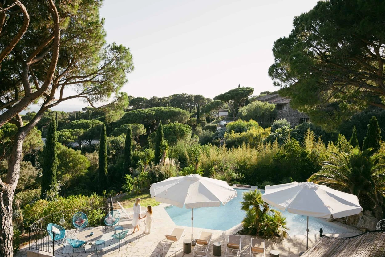 exterieur-d-une-villa-entoure-de-verdure-avec-deux-parasols-blancs-quatre-chaises-longues-cinq-chaises-bleu-une-maman-et-sa-fille-autour-de-la-piscine
