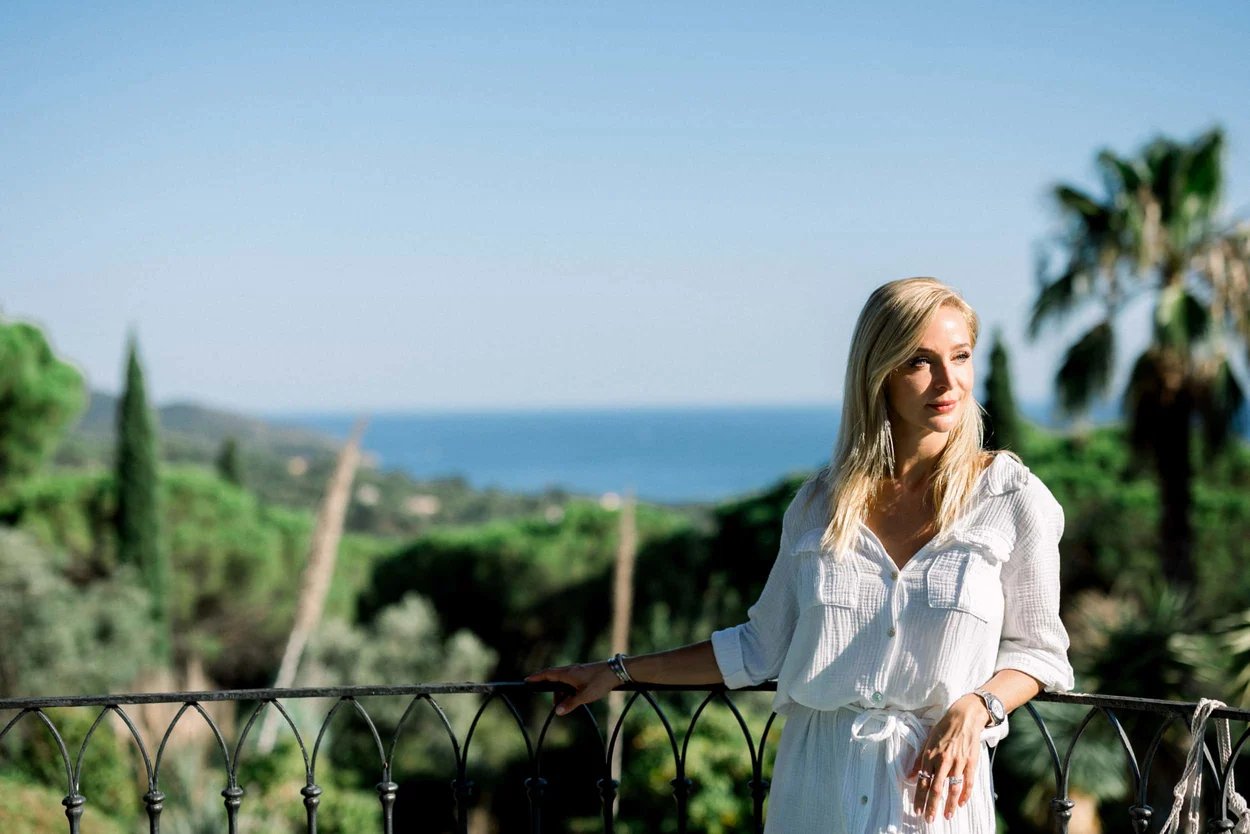 femme-blonde-habillee-en-blanc-sur-le-balcon-d-une-villa-regardant-la-vue-sur-la-verdure-et-la-mer-en-arriere-plan
