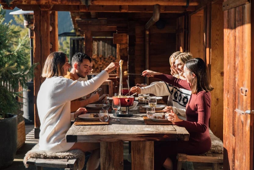 six-amis-femmes-et-homme-assis-autour-d-une-table-en-bois-sur-la-terrasse-d-un-chalet-face-a-la-foret-partageant-une-fondue-dans-un-plat-rouge