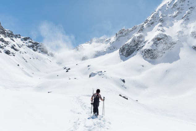homme-brun-de-dos-avec-une-salopette-noire-et-un-pull-a-rayures-tenant-une-paire-de-ski-marchant-vers-les-montagnes-enneigees-sous-le-ciel-bleu