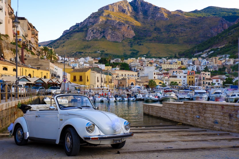 voiture-de-collection-grise-cabriolet-garee-sur-des-paves-devant-un-port-en-sicile-avec-des-bateaux-un-village-et-la-montagne-en-fond