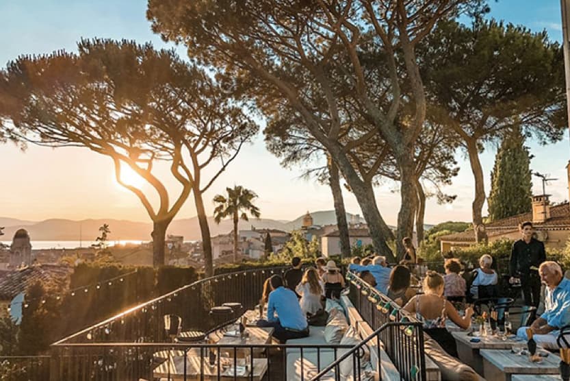 terrasse-du-rooftop-le-tigrr-saint-tropez-clients-assis-autour-des-tables-sous-les-arbres-devant-une-vue-sur-la-ville-etla-mer-au-loin