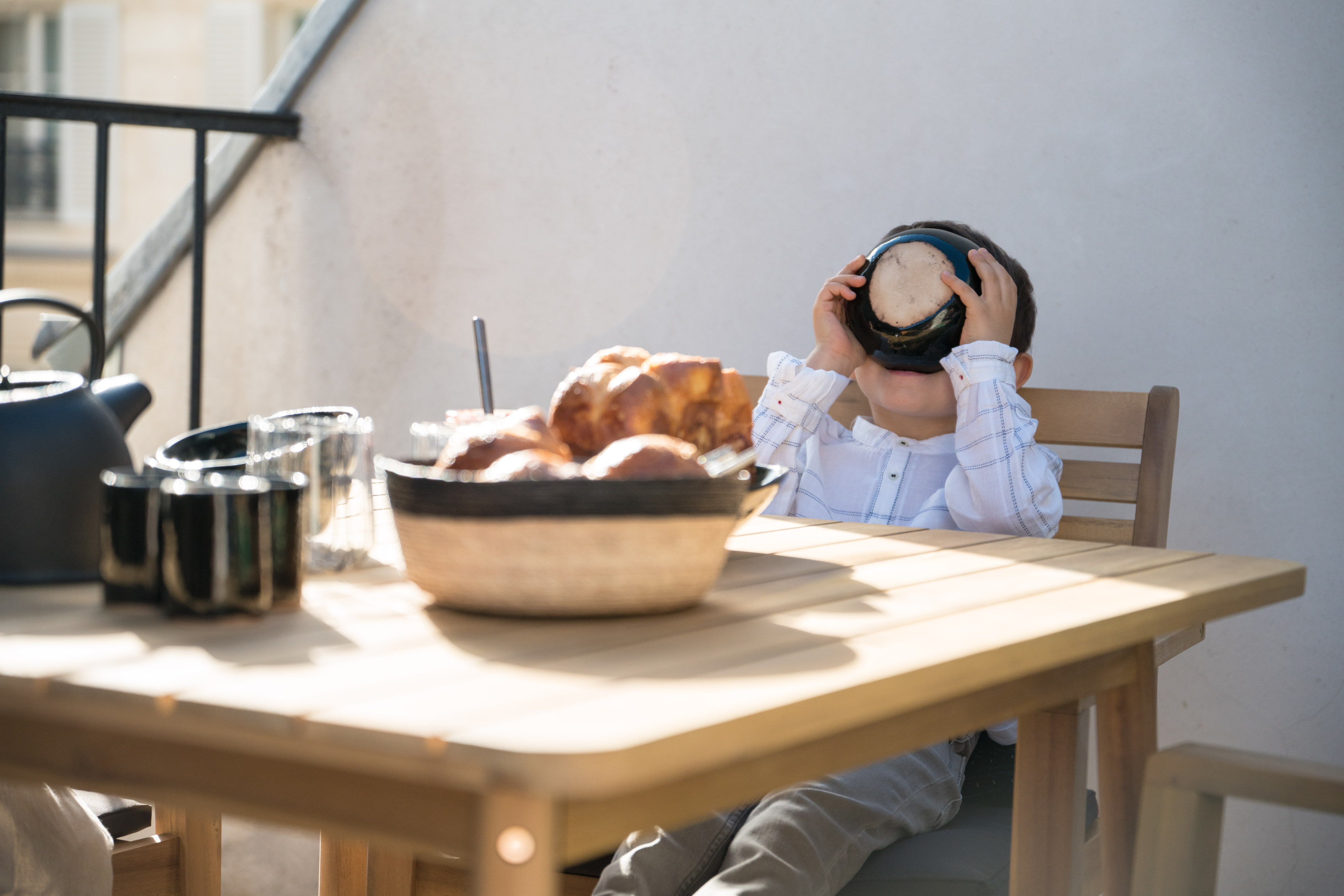 petit-garcon-en-chemise-blanche-qui-boit-dans-un-bol-noir-assis-sur-une-chaise-en-bois-devant-une-table-en-bois-remplie-de-viennoiseries