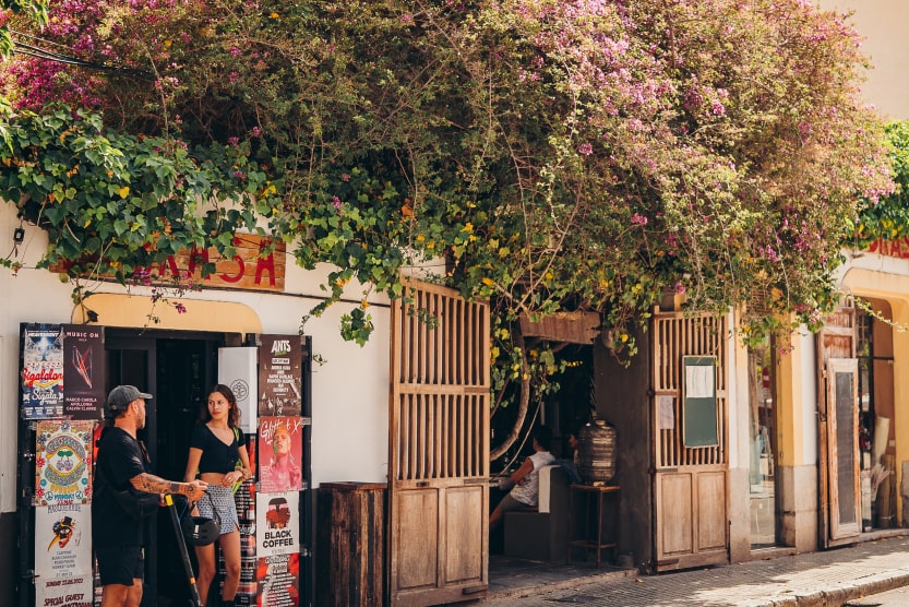 un-homme-en-trottinette-electrique-discute-avec-une-femme-devant-une-petite-boutique-avec-des-posters-de-festivals-dans-une-ruelle-fleurie-de-eivissa