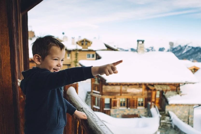 petit-garcon-brun-en-pull-bleu-pointant-quelque-chose-du-doigt-debout-sur-le-balcon-d-un-chalet-face-a-un-village-alpin-recouvert-de-neige