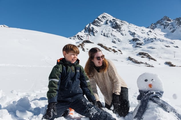 a boy-in-skiing-suit-and-a-woman-smiling-to-the-side-with-a-snowman-with-a-scarf-in-front-of-the-mountains