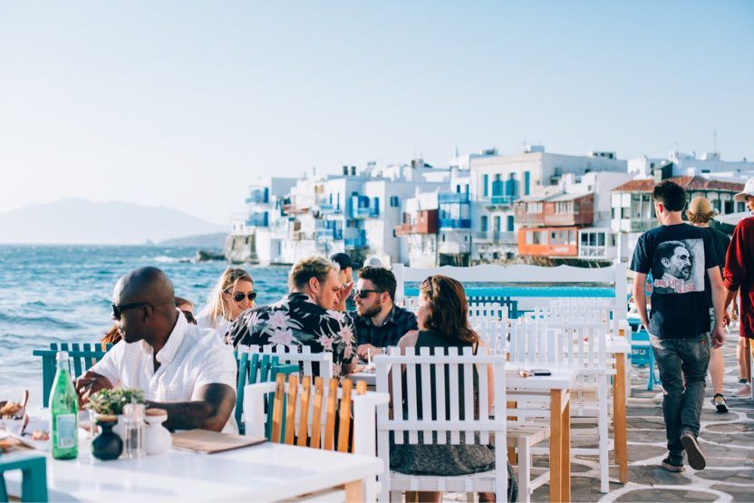groupe-d-amis-assis-sur-la-terrasse-d-un-restaurant-sur-le-port-de-mykonos-face-a-la-mer-egee-et-au-village-avec-des-maisons-colorees