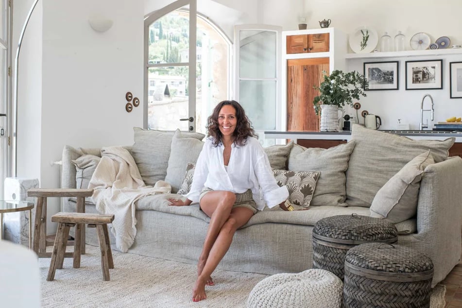 femme-brune-en-chemise-et-short-souriante-assise-sur-un-canape-beige-dans-un-salon-a-la-decoration-beige-et-en-bois