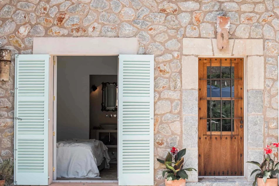 vue-sur-la-porte-fenetre-d-une-chambre-avec-des-volets-bleus-a-cote-d-une-porte-en-bois-entoure-de-deux-pots-de-fleurs-roses-sur-une-maison-en-pierre