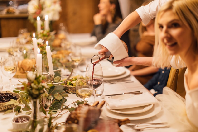 une-femme-blonde-assise-a-une-grande-table-de-noel-decoree-avec-des-plantes-et-des-bougies-se-fait-servir-du-vin-rouge-dans-son-verre